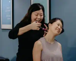 A female chiropractic practitioner using a handheld instrument-assisted adjustment tool on the side of a female patient's neck. The patient is seated, smiling, and tilting her head to the side during the treatment.