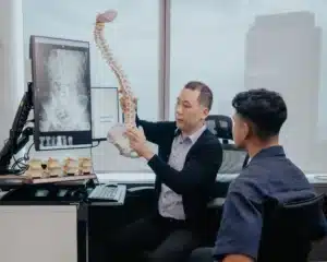 A chiropractor in an office consulting with a male patient. The practitioner is holding and pointing to a flexible anatomical model of a human spine and pelvis, while a large vertical monitor next to him displays an X-ray of a lumbar spine.
