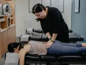 A female chiropractor performing a manual adjustment on a patient's lower back and pelvis area while the patient lies face down on a chiropractic table.