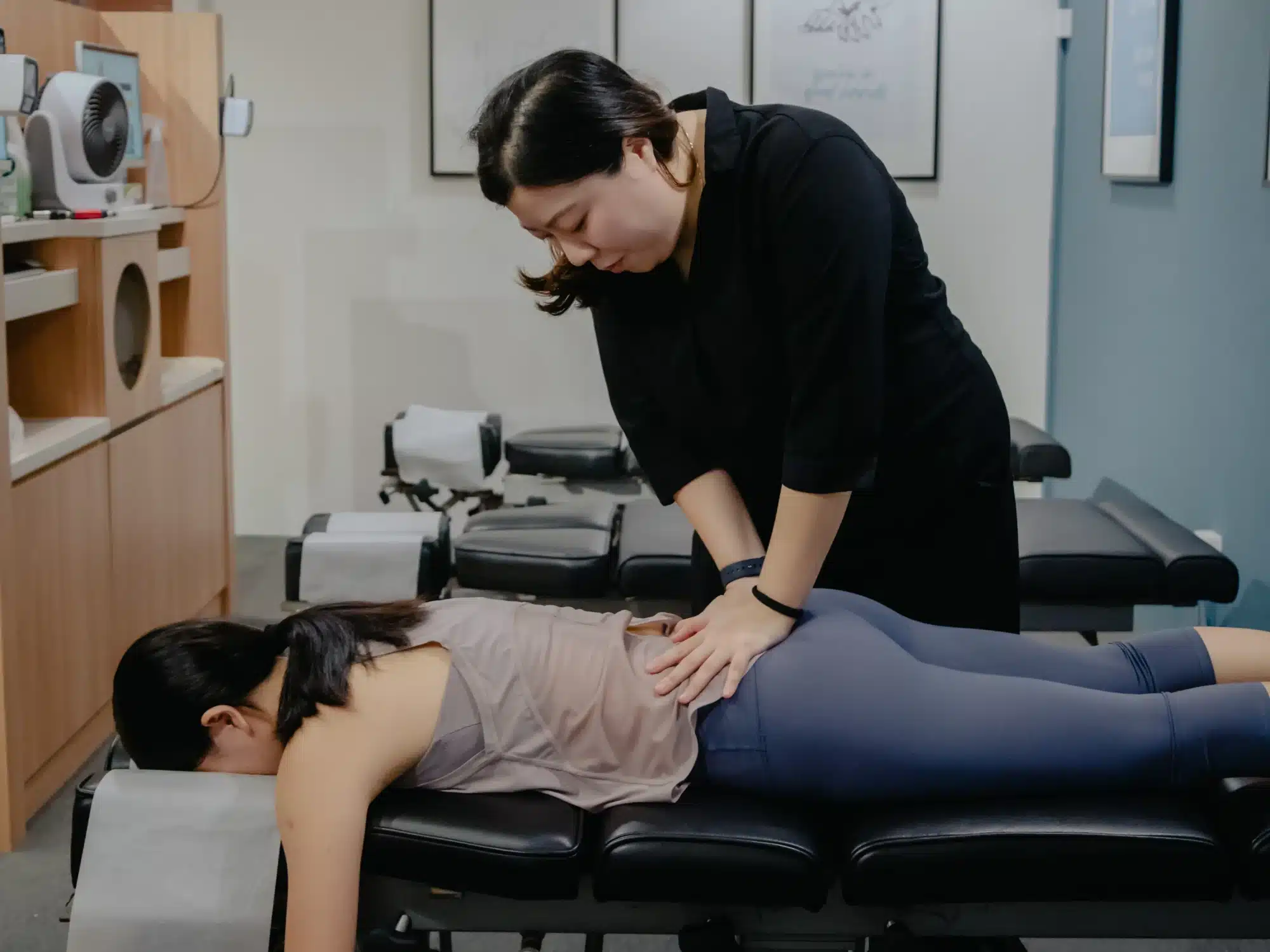 A female chiropractor performing a manual adjustment on a patient's lower back and pelvis area while the patient lies face down on a chiropractic table.