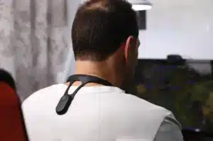 Rear view of a man sitting at a computer desk wearing a small, electronic smart posture trainer device around his neck and upper back.