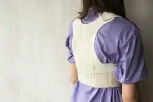 Rear view of a person wearing a beige fabric medical posture support vest over a purple t-shirt.