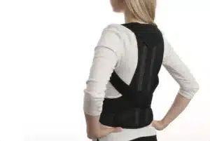 Rear view of a woman wearing a black full-back posture corrector brace over a light-coloured long-sleeve shirt.