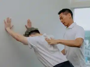 A practitioner guides a patient's posture as the patient leans forward with hands pressed flat against a white wall for a stretch.